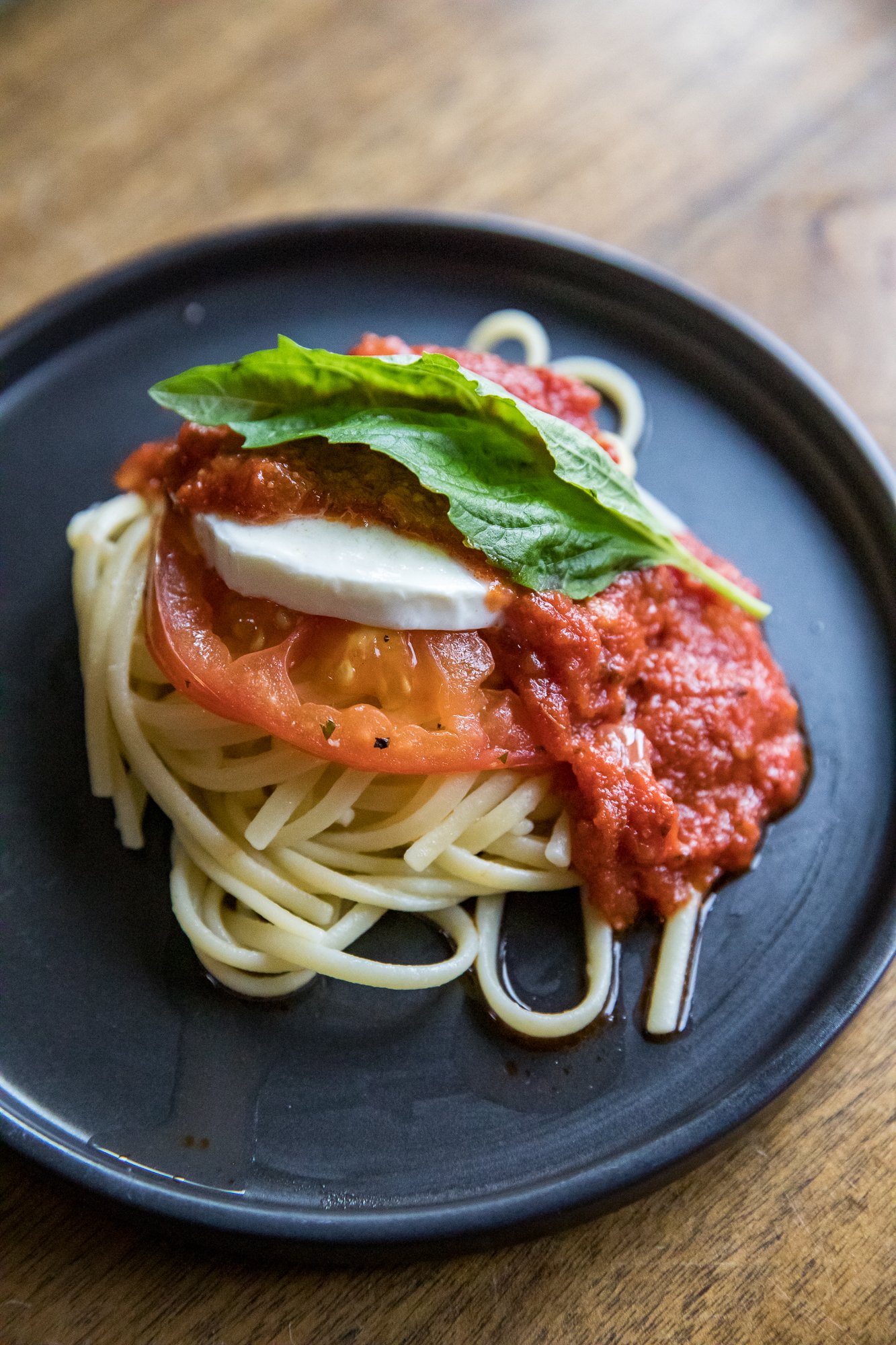 Caprese Pasta Marinara on a black plate sitting on a brown wood table with basil and fresh mozzarella on top